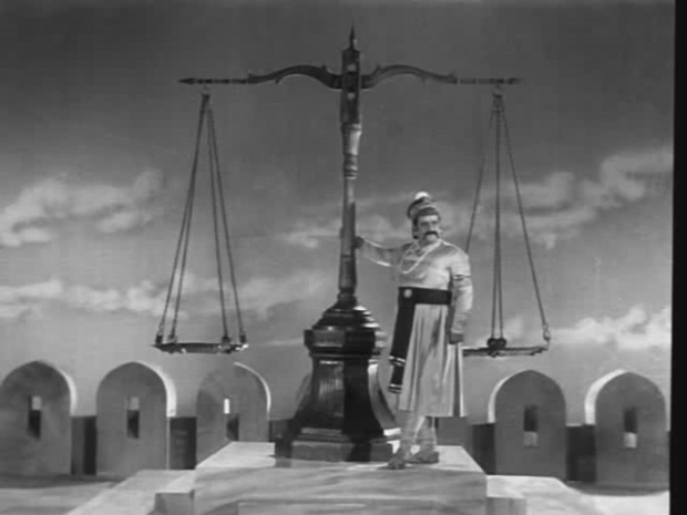 black and white film still showing a tall man in traditional south asian dress resting against a human sized set of weighing scales