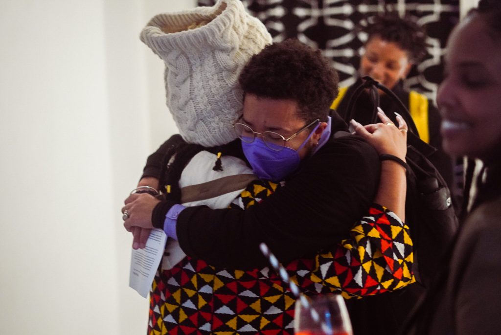 HCJ wearing a woolen crown and colourful robe embraces a visitor who is wearing a mask over their nose and mouth.