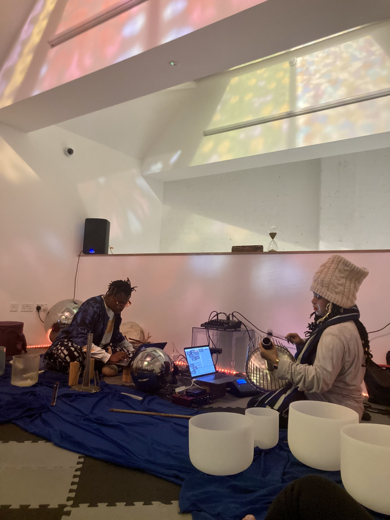 Evan Ifekoya and HCJ perform together. They are sat on the floor surrounded by singing bowls, waterphones, percussion and mirror balls. The stained glass above creates colourful shadows.